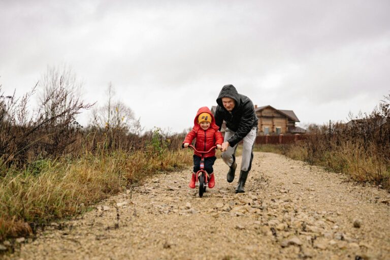 A father helps his child ride a bicycle on a rural dirt path, showcasing bonding and joy.