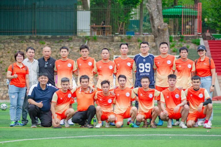 Outdoor group photo of a Vietnamese soccer team in orange uniforms at a park in Hanoi.