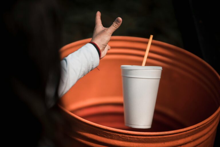 A hand aiming to throw a white disposable cup into an orange garbage bin, emphasizing waste disposal.