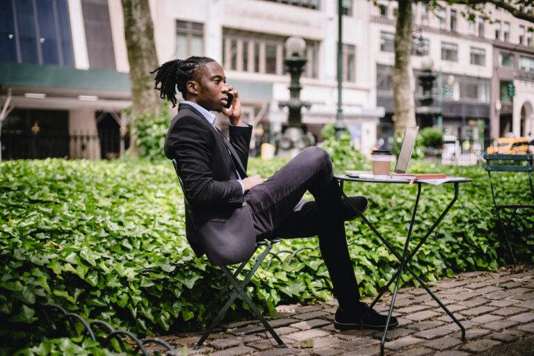 Side view serious African American businessman in black formal wear sitting at table in city park with legs crossed and having conversation via modern smartphone