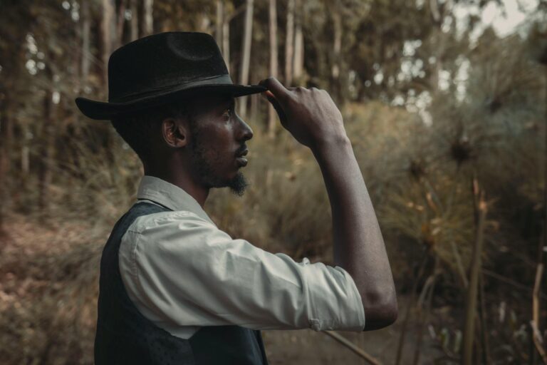 Side view of a stylish man wearing a hat and vest in a forest setting.