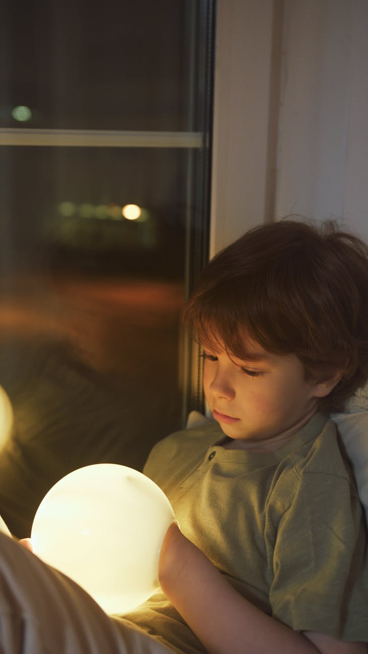 A child sitting indoors holding a glowing ball by a window at night.