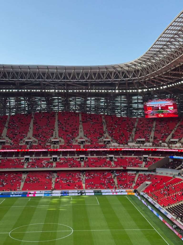 A lively football stadium with red seats and spectators under a clear blue sky.