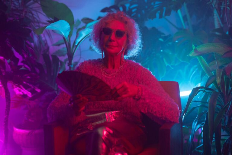Elderly woman with sunglasses in a chic, neon-lit interior, exuding style and confidence.
