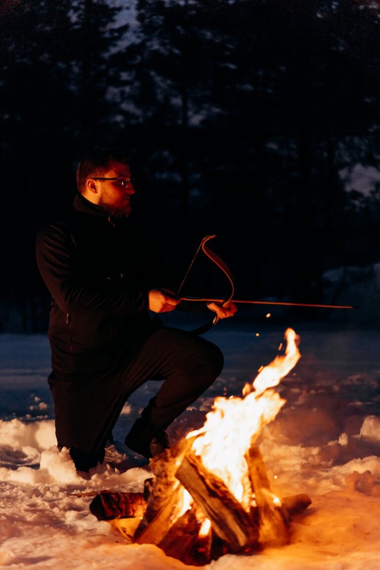 A man enjoying a winter night by the campfire with his bow and arrow in the snowy outdoors.