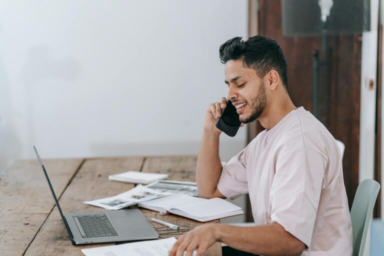 Young Hispanic man engaged in a phone call while working remotely on a laptop at home.