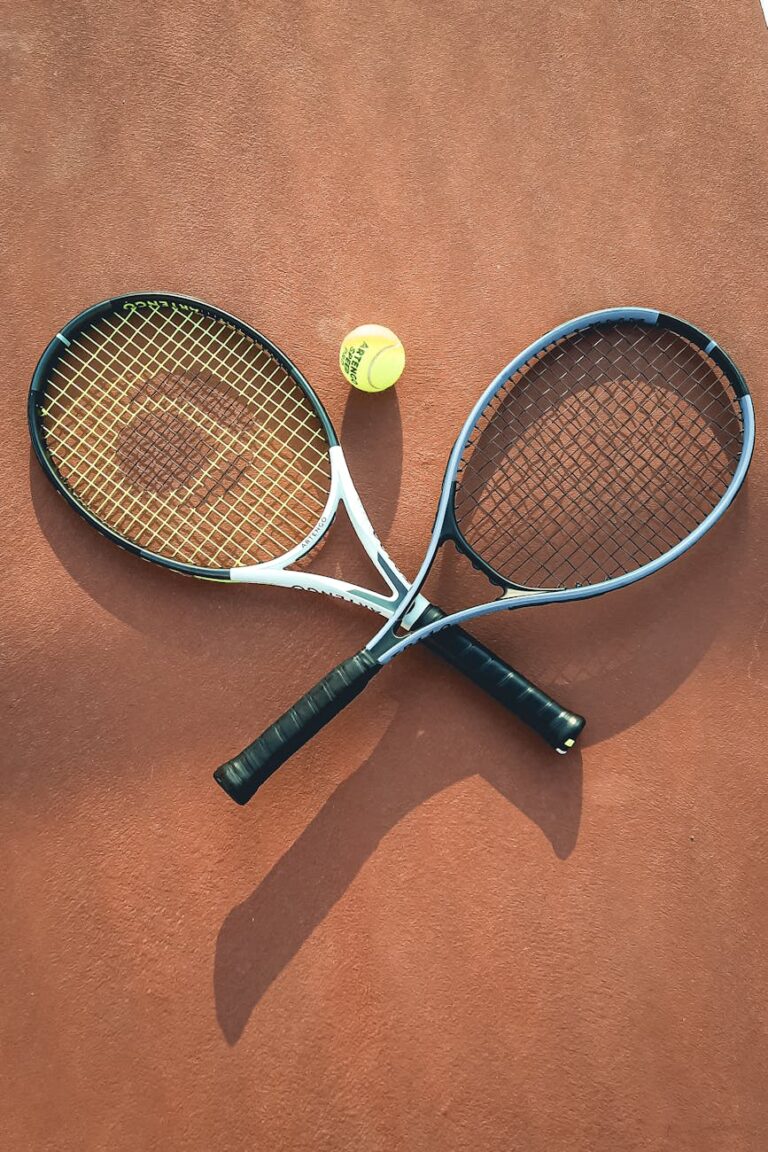 Two tennis rackets and a ball arranged on a clay court, casting shadows.