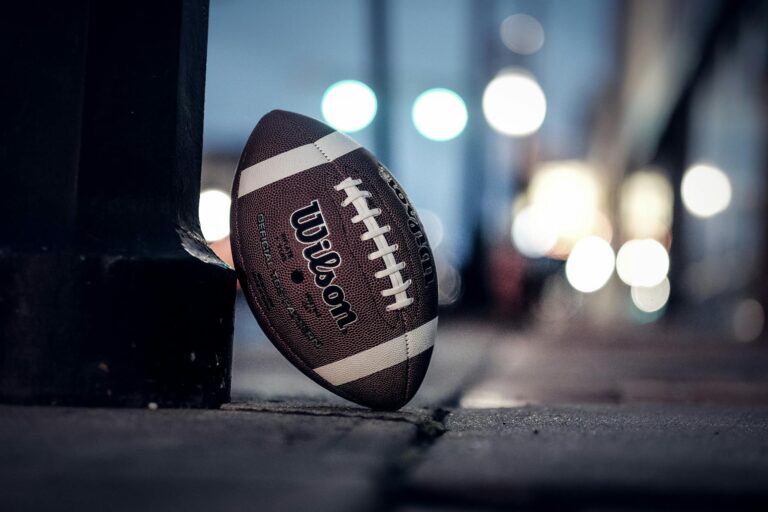 A Wilson American football rests on a city sidewalk at night with bokeh lights in the background.