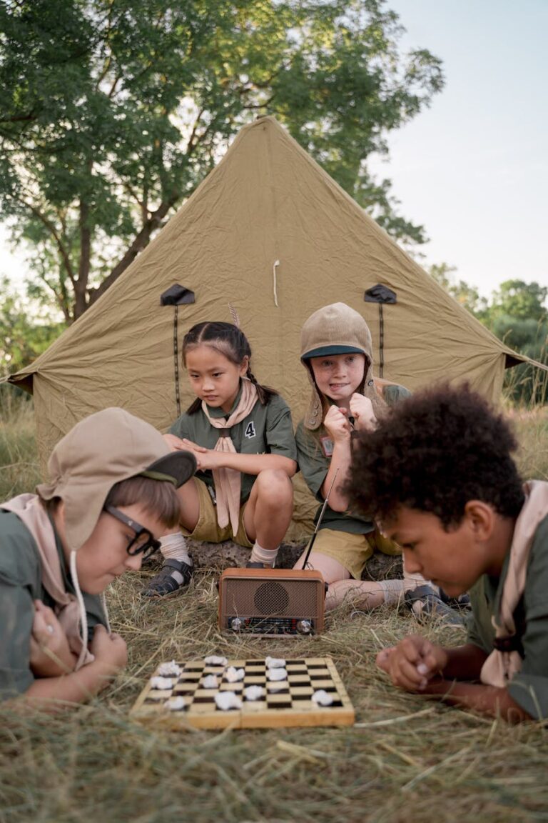 Group of diverse children playing chess outdoors in front of a tent at a scout camp.