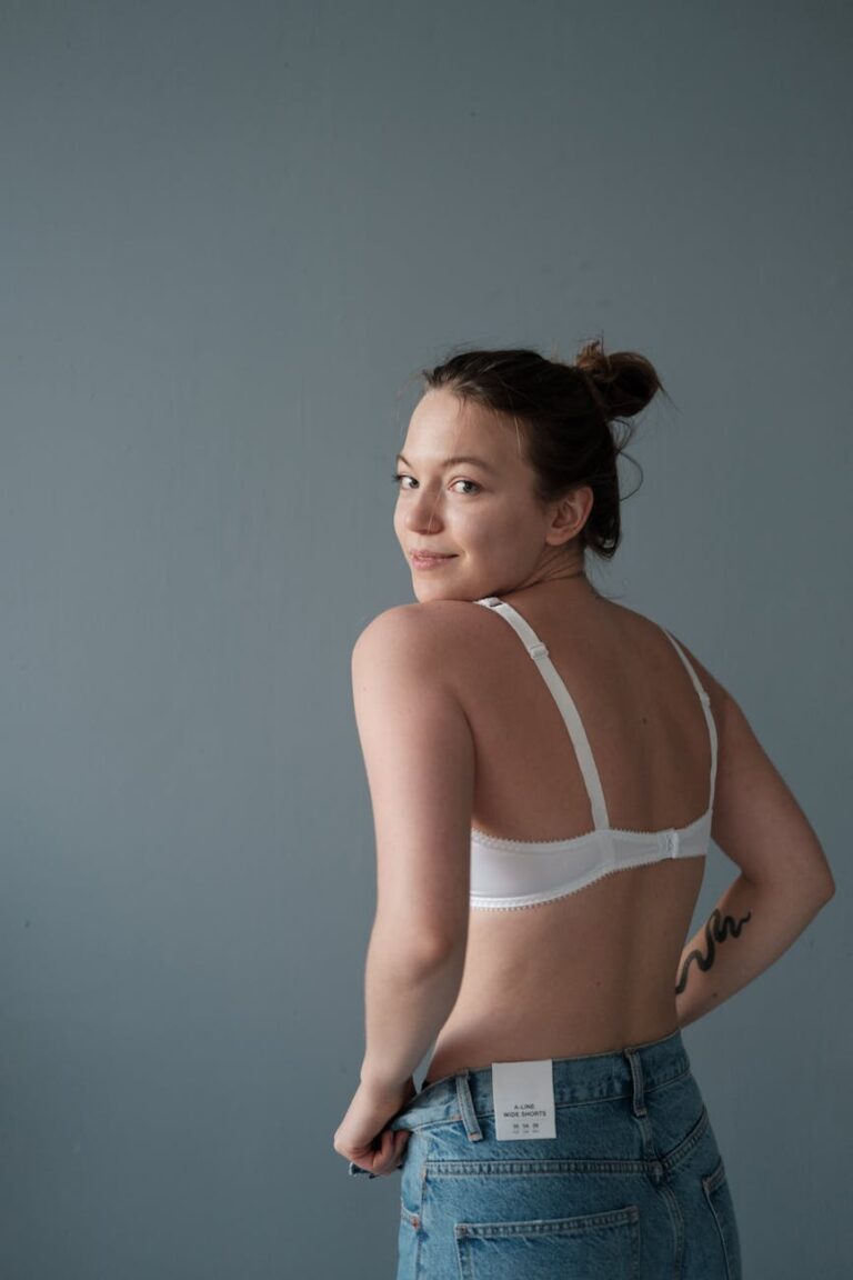 Caucasian woman posing confidently in denim jeans and white bra with a minimalist backdrop.