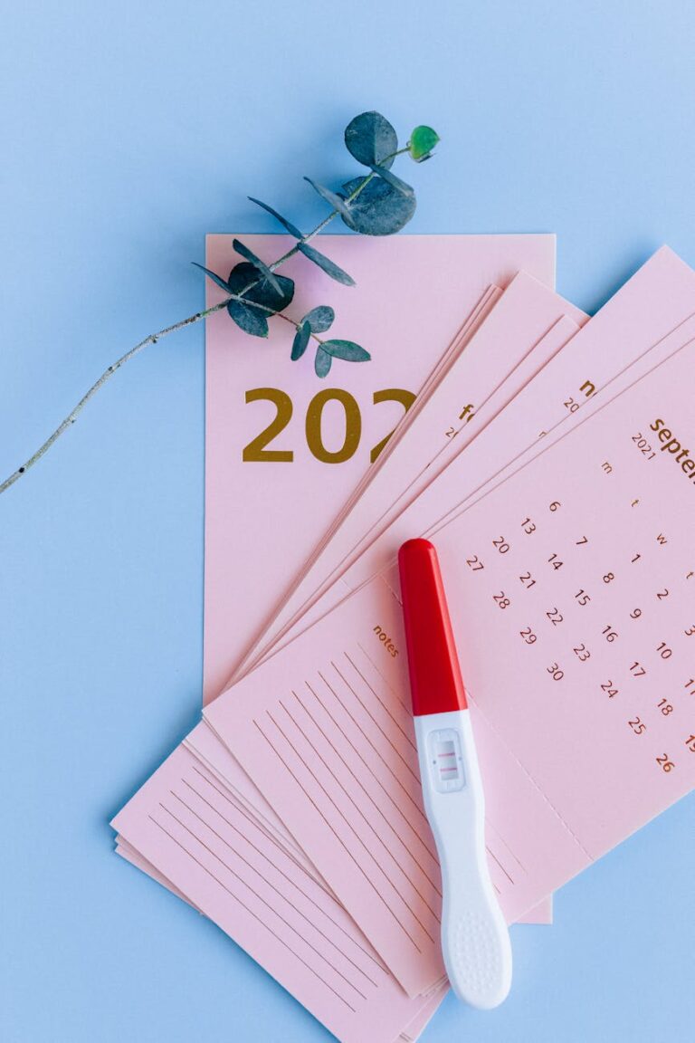 Pregnancy test atop a pink calendar with eucalyptus on a blue surface, symbolizing anticipation.