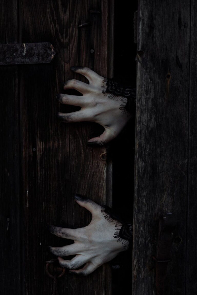 Eerie white hands with black nails reach through a dark wooden door, perfect for Halloween themes.