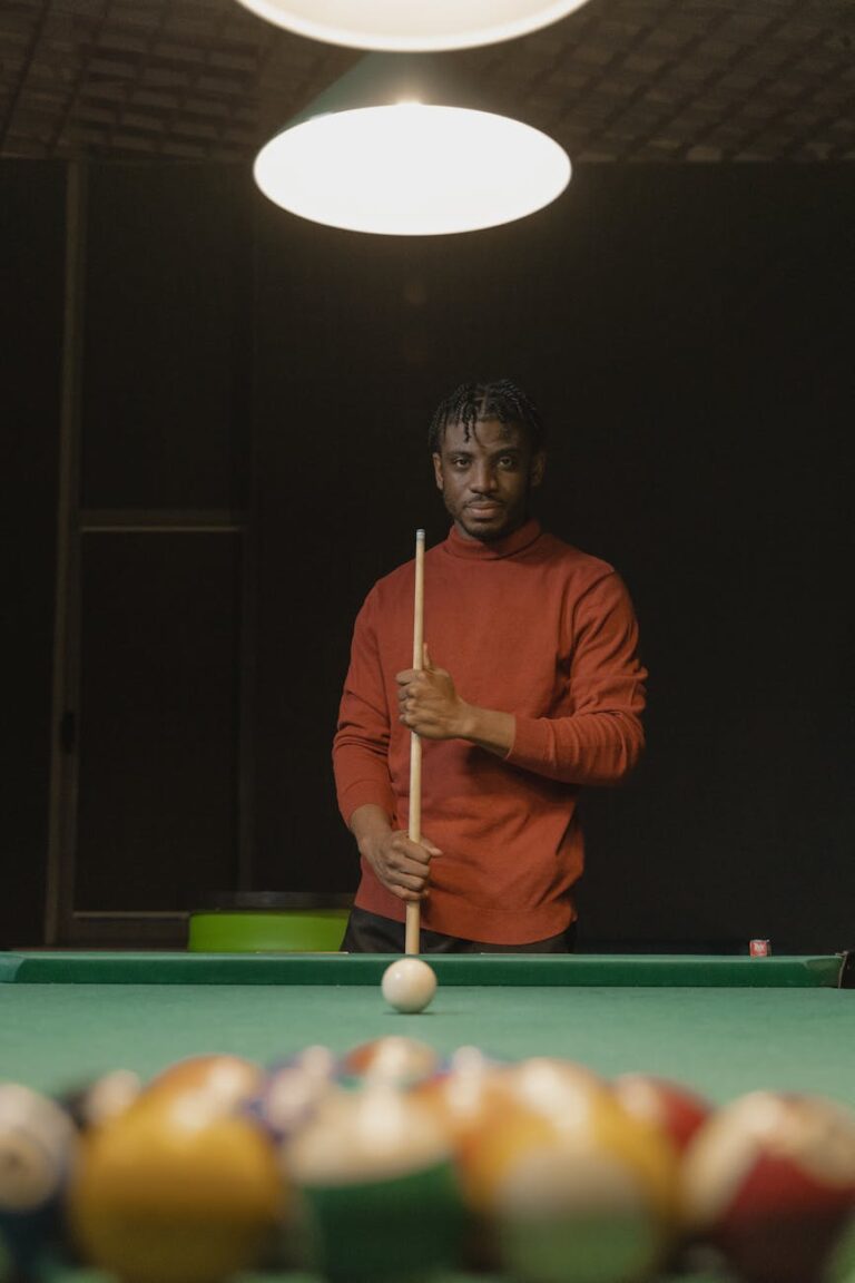 A young man in a red sweater holds a cue stick at a pool table, focused and poised.