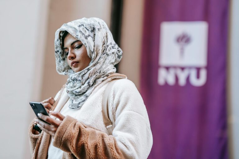 A young woman wearing a hijab stands in front of NYU, using her smartphone outdoors.