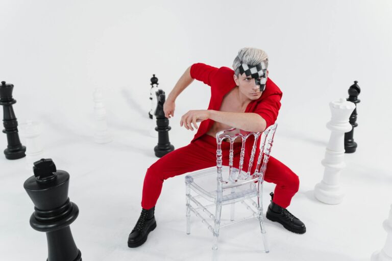 Man in red suit with chess-themed eye mask surrounded by large chess pieces.