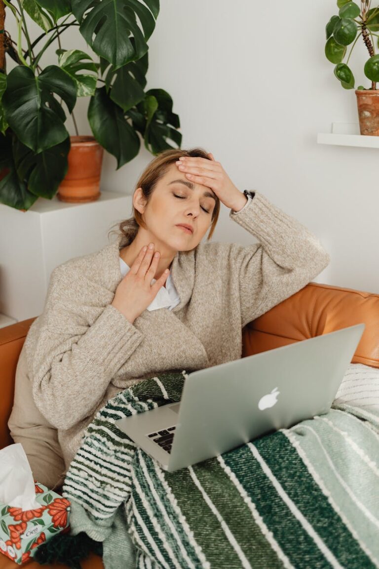 Woman with fever using a laptop while wrapped in a blanket at home, resting on a sofa.