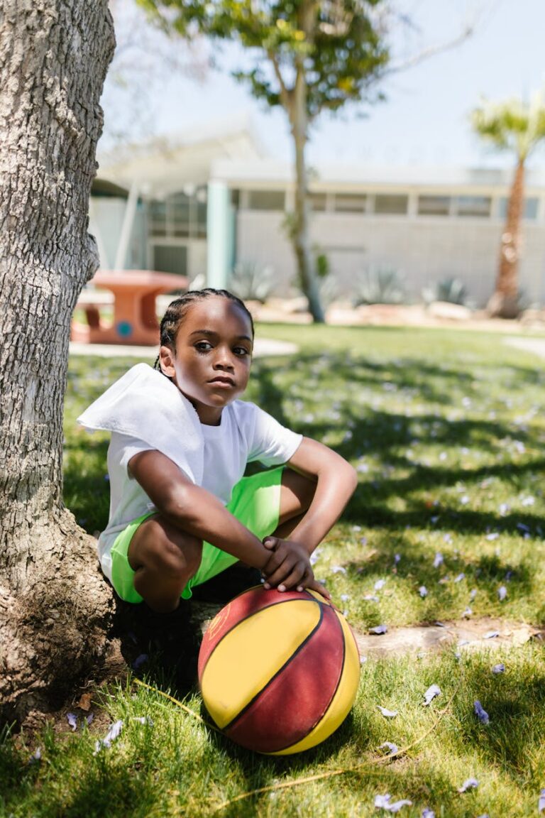 A young boy sits in a park holding a basketball on a sunny day.