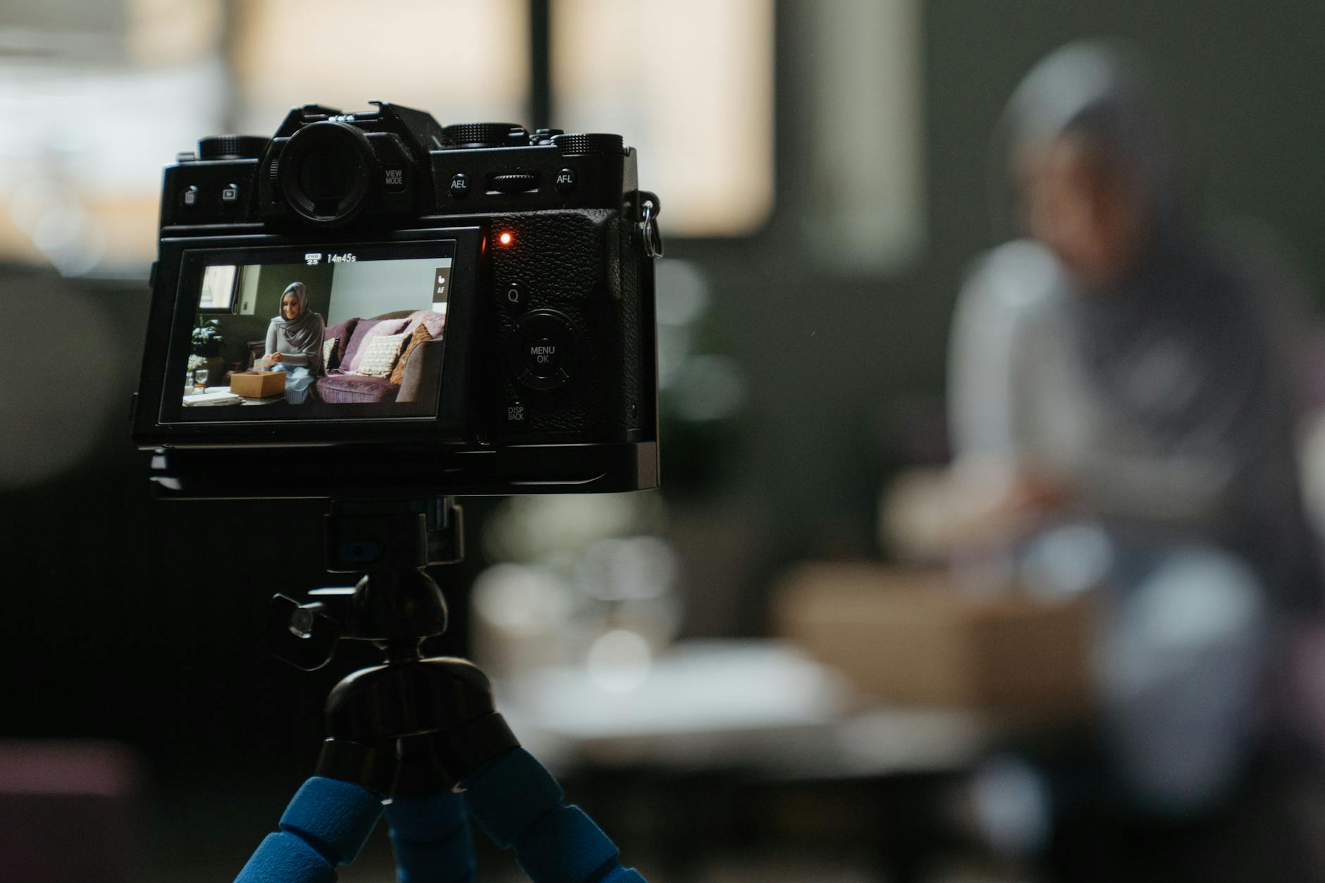 A vlogger sits in a living room setting while being recorded by a mirrorless camera on a tripod.
