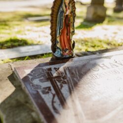 Statue of Virgin Mary on a gravestone in a sunlit cemetery, symbolizing spirituality and remembrance.