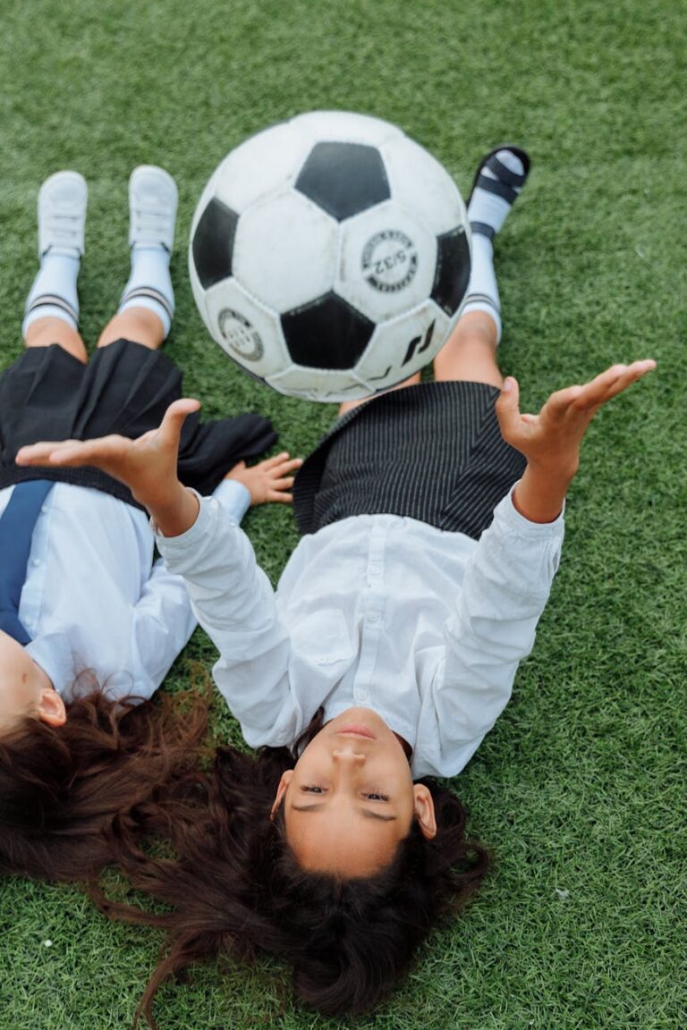 Two girls lying on grass, joyfully playing with a soccer ball outdoors.