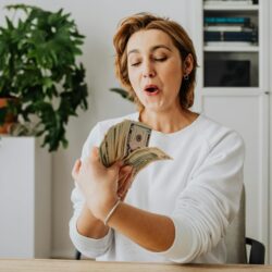 A woman in a white sweater excitedly counts American dollar bills indoors, reflecting joy and prosperity.