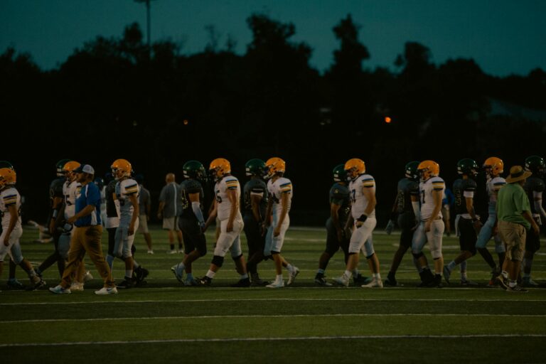 Teams shake hands post-game on a football field under the night sky.