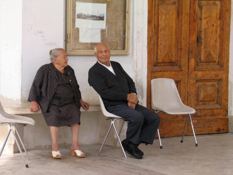 Elderly couple sitting on chairs outside an Italian building, conversing.