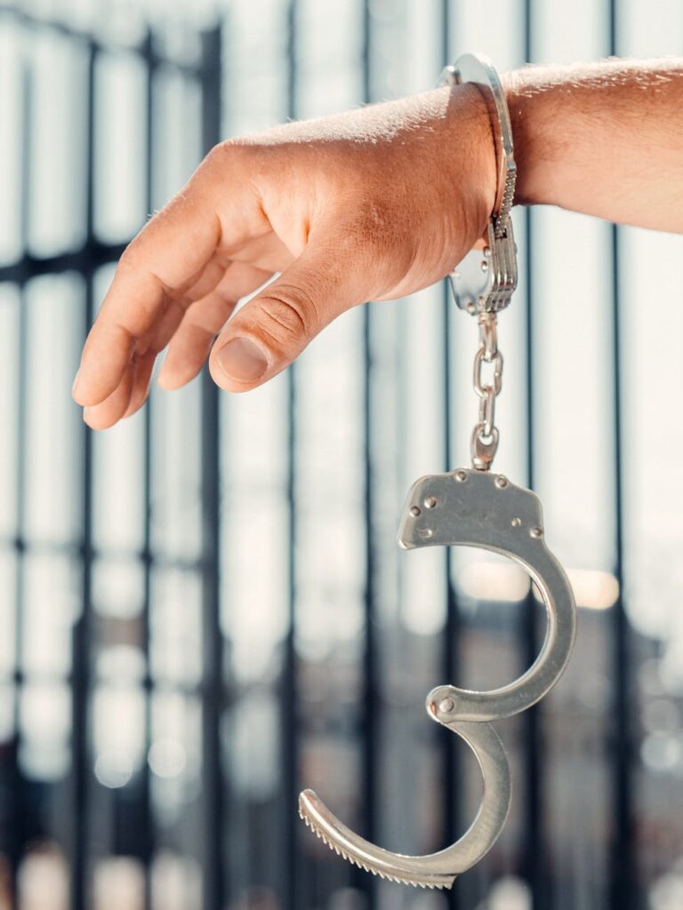 A close-up, conceptual shot of a handcuffed hand with one open handcuff against blurred bars.