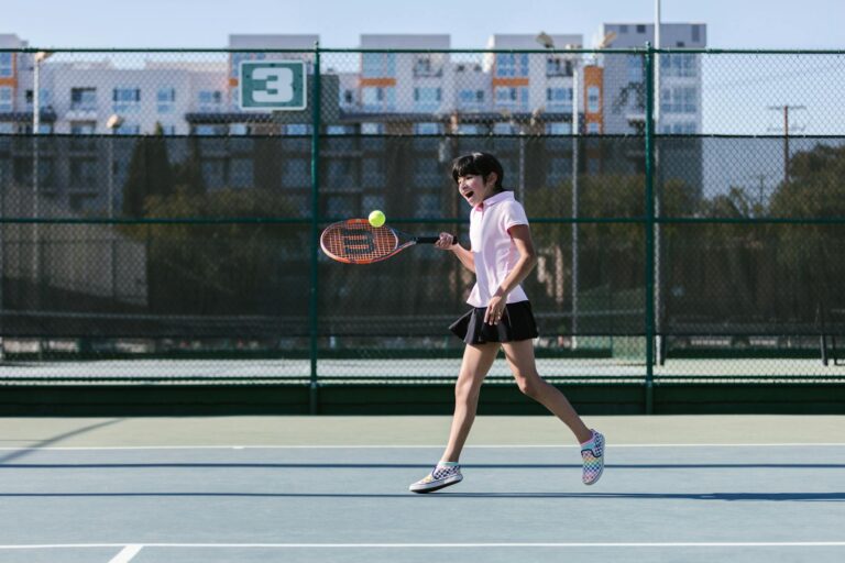 Young girl joyfully hitting a tennis ball on an outdoor court during a sunny day.