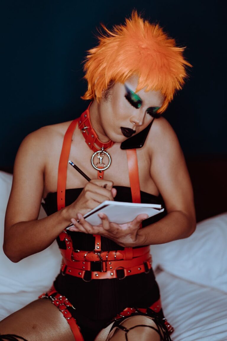 Serious young ethnic androgynous guy with aggressive makeup in provocative BDSM costume and orange wig sitting on bed and taking notes in diary during phone conversation