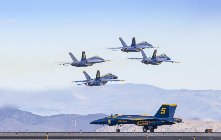 U.S. Navy Blue Angels jets in formation flying over a runway with mountains in the background.