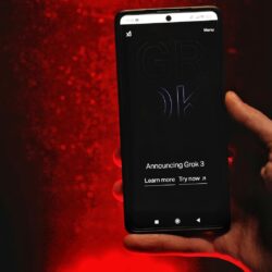 A hand holds a smartphone displaying Grok 3 announcement against a red background.