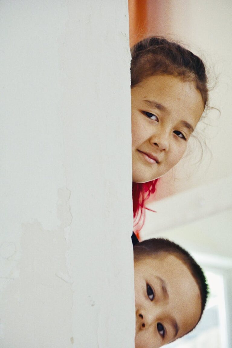 Two children peeking from behind a white wall indoors in Talgar, Kazakhstan.
