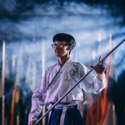 A man in traditional clothing performs with a staff at a night ceremony with flags in the background.