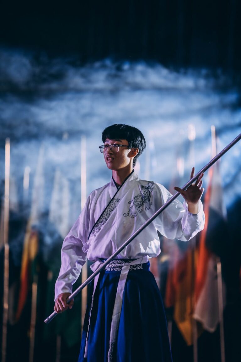 A man in traditional clothing performs with a staff at a night ceremony with flags in the background.