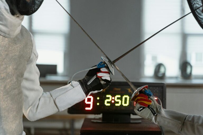 Two fencers in action, crossing foil swords in an indoor arena with a timer in the background.