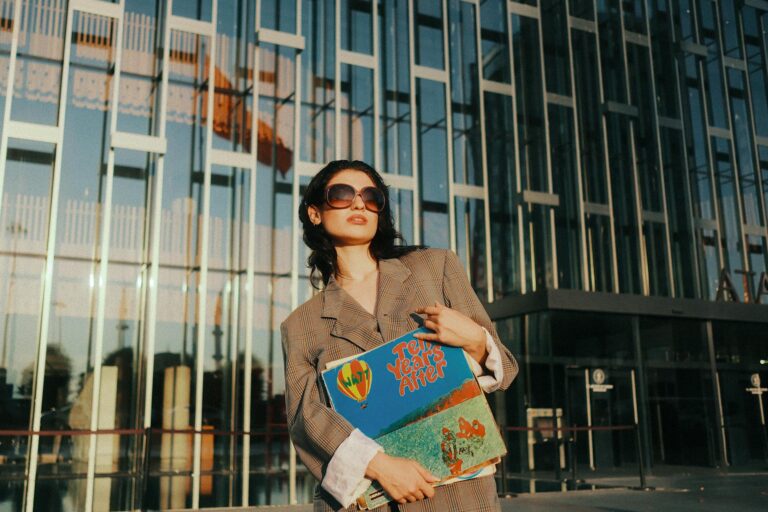 Fashionable woman in sunglasses holding vinyl records outside modern building.