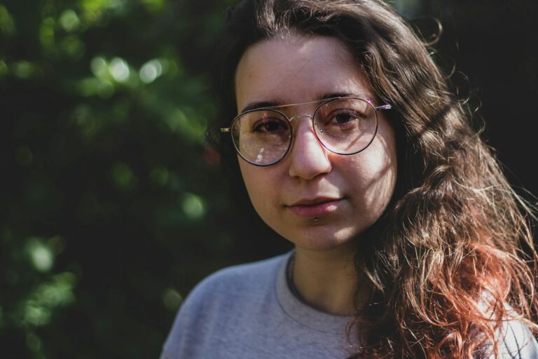 Close-up portrait of a woman with glasses and long hair, set outdoors with natural lighting.