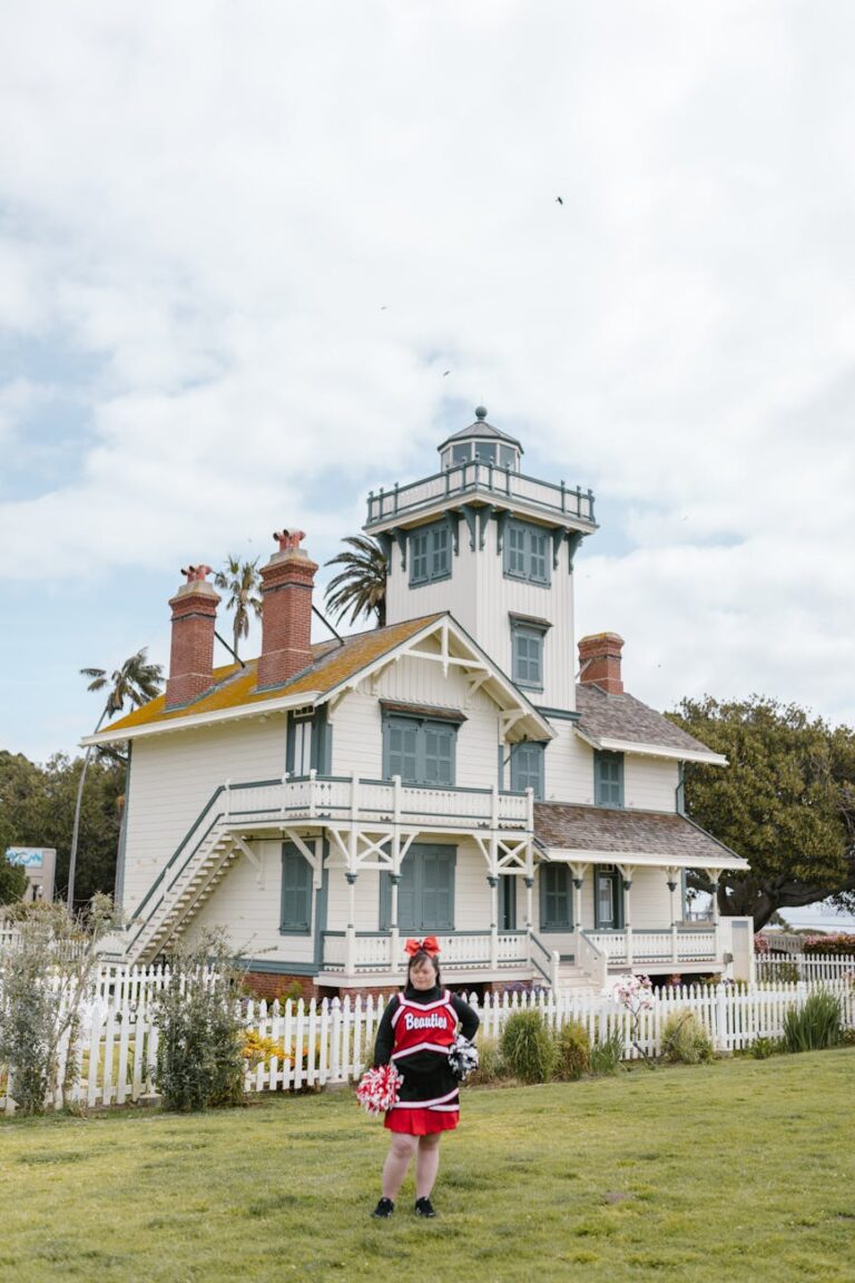A cheerleader poses confidently with pompoms in front of a historic lighthouse on a bright day.