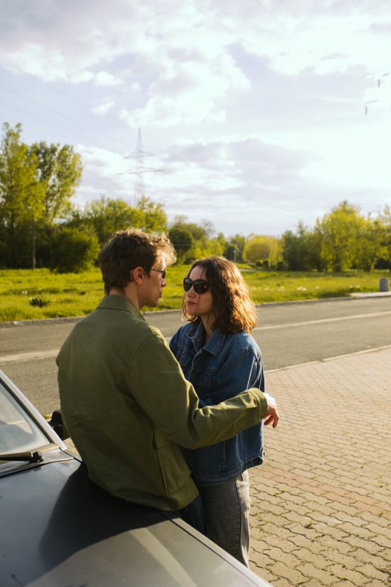 Young couple outdoors, leaning on a car, embracing and enjoying a sunny day.