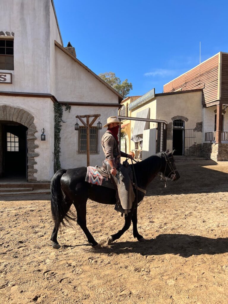 Cowboy in traditional attire rides black horse through historic Western town.