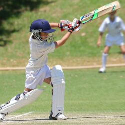 Young cricketer in white gear executing a sweep shot on a sunny day.