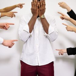 A man in distress covers his face surrounded by multiple pointing fingers.