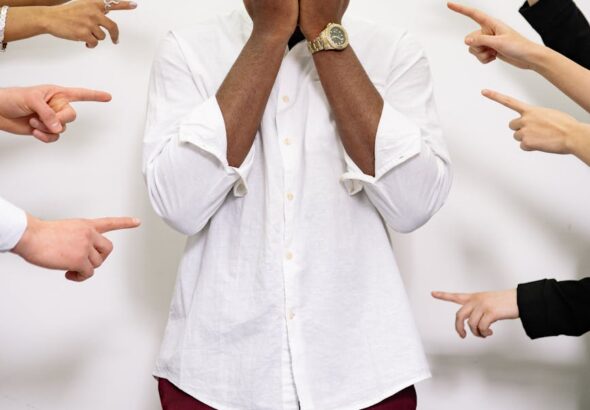 A man in distress covers his face surrounded by multiple pointing fingers.