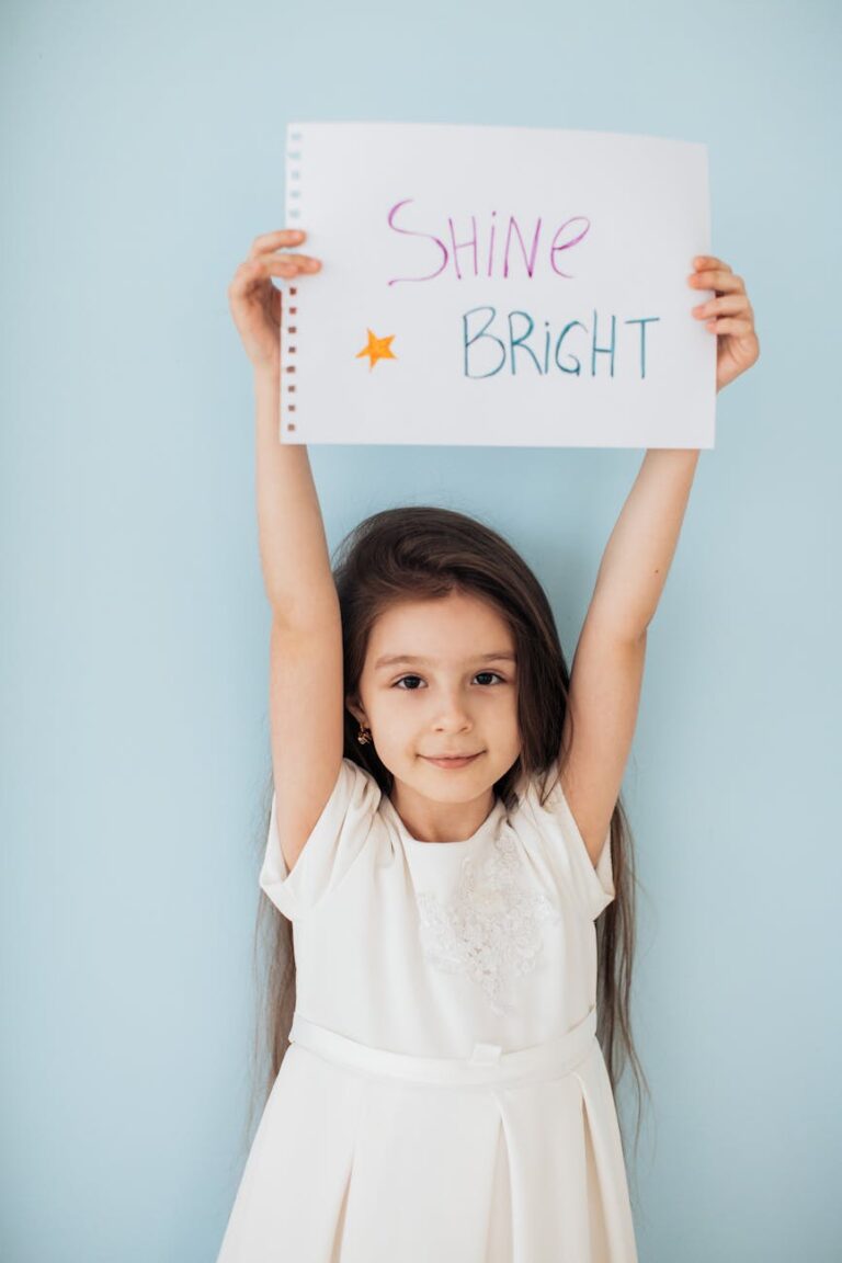 Cute girl in a white dress holds a 'Shine Bright' sign against a light blue background.