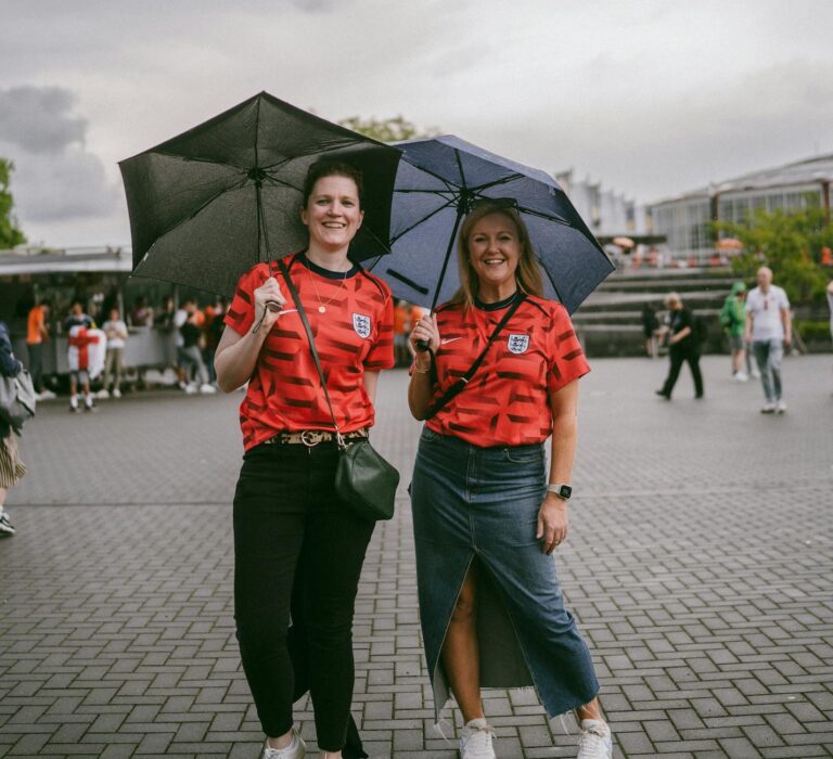Two women in red jerseys standing with umbrellas, smiling at an outdoor sports event.