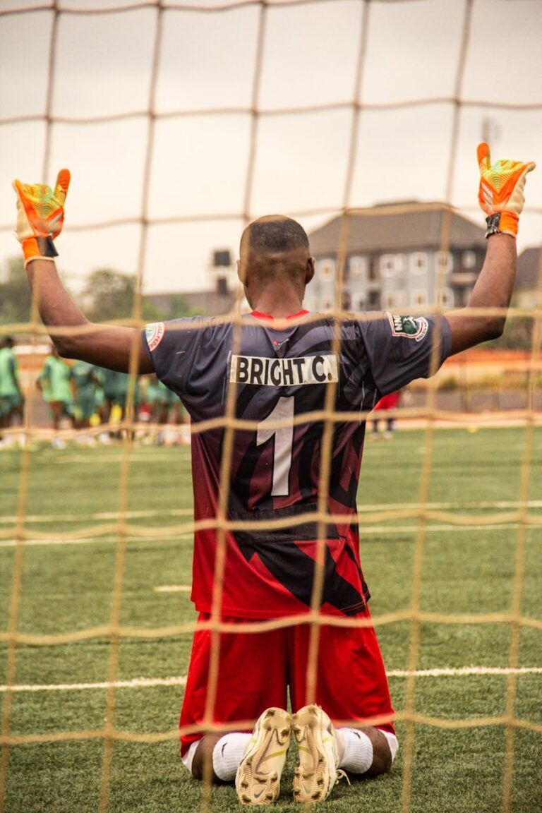 Soccer goalkeeper kneeling on the grass field with gloves raised in celebration.