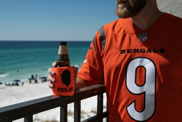 Man in Bengals jersey enjoying beer by the beach on a sunny day.