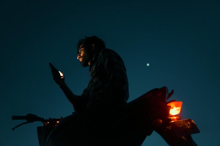 Silhouetted man on motorcycle using smartphone under night sky with star visible.
