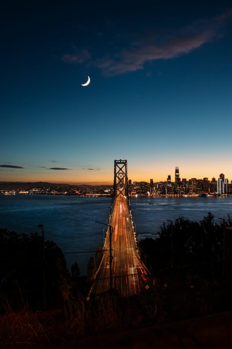 Dramatic night view of the Bay Bridge with city skyline and crescent moon under a starry sky.